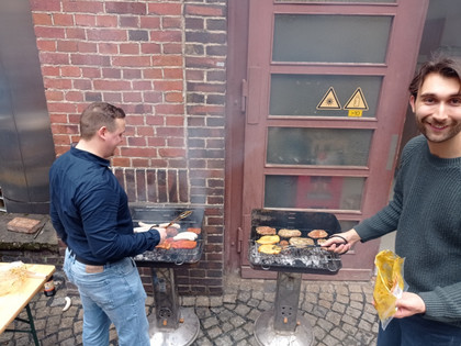 Zwei Männer stehen vor Holzkohlegrills im Innenhof, einer grillt Burger und Würstchen, der andere lächelt in die Kamera und hält Grillzange und Packung