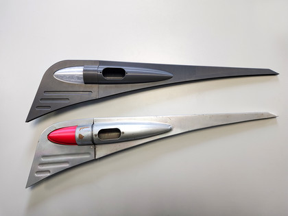 Zwei metallische Flugzeug-Bauteile in Form aerodynamischer Finnen liegen nebeneinander auf einem hellen Tisch; das obere Bauteil ist dunkelgrau, das untere silbern mit roter Spitze.