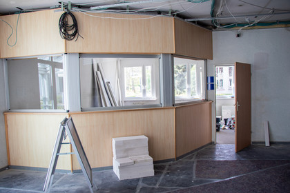 Innenaufnahme eines Gebäudes im Umbau, zeigt eine halbhohe Holzverkleidung mit Fenstern, dahinter ein Raum mit offen stehender Tür, auf dem Boden liegen Baustellenmaterialien und eine Leiter, Kabel hängen von der Decke.