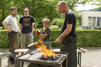 **Alt-Text:** Ein Junge steht mit Handschuhen vor einer Schmiedestelle und wird von einem Schmied mit Lederschürze angeleitet; zwei weitere Männer beobachten die Szene im Hintergrund, im Vordergrund lodert ein Kohlefeuer.