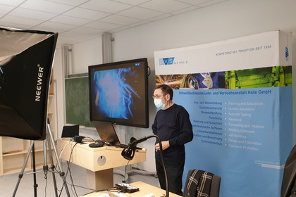 Ein Mann mit Mundschutz steht in einem Präsentationsraum vor einem Banner der SLV Halle GmbH, auf einem Bildschirm ist ein Lichtbogen-Schweißprozess zu sehen, Kameratechnik und Laptops sind im Vordergrund aufgebaut.
