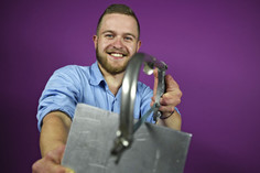 Ein junger Mann mit Vollbart und blauem Hemd lächelt in die Kamera und hält ein geschweißtes Metallbauteil mit gebogenem Rohr vor einem lila Hintergrund.