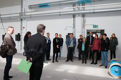 Rund 15 Personen stehen in einer Industriehalle mit sichtbaren Lüftungsrohren, ein Mann im Anzug mit grünem Ordner spricht zur Gruppe, Tageslicht fällt durch ein Fenster