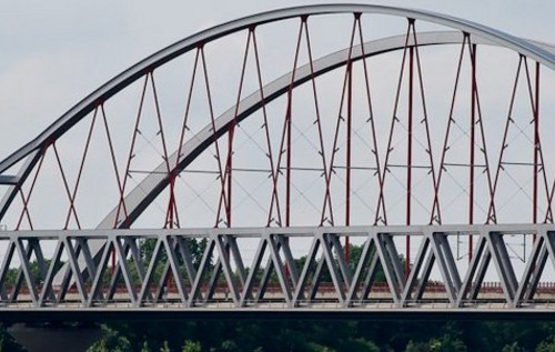 Stahlbrücke mit bogenförmiger Tragkonstruktion und Fachwerkträgern über einem Gewässer vor bewölktem Himmel