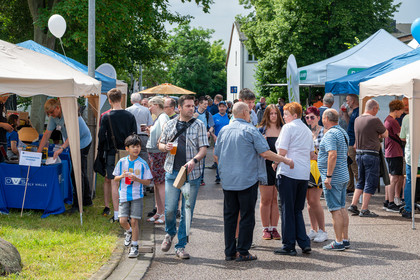 Der Weg zwischen den Gebäuden der SLV Halle ist voller Menschen, die sich unterhalten. An den Seitenrändern sind weiße Pavillons aufgebaut und im Hintergrund sind Luftballonns zu sehen. Es herrscht eine entspannte Atmosphäre bei Sonnenschein.