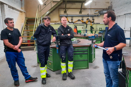 Vier Männer stehen in einer Werkhalle um eine grüne Werkbank und sprechen miteinander. Drei tragen dunkle Arbeitskleidung, zwei davon mit auffälligen gelben Reflexbereichen an den Hosen, im Hintergrund sind Werkzeuge, Maschinen und eine Treppe zu sehen.
