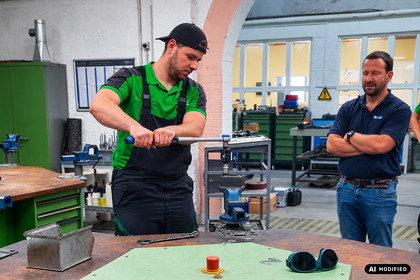 Zwei Männer stehen an einem Arbeitsplatz in einer Werkhalle. Einer bearbeitet ein eingespanntes Metallbauteil mit einem Handwerkzeug, während der andere mit verschränkten Armen danebensteht und zusieht.