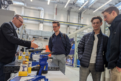 Vier Männer in technischer Werkhalle, ein Mann im Anzug erklärt Werkzeuge auf einem Tisch, darunter blaue Nietgeräte und Verpackungen der Marke GESIPA