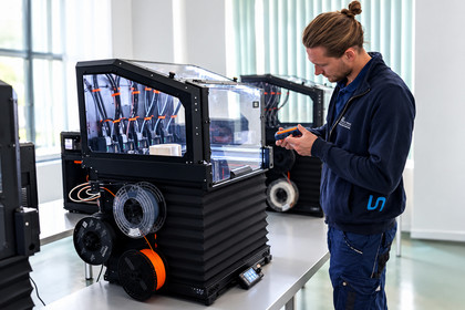 Ein Mann in blauer Arbeitskleidung betrachtet ein blau-oranges Bauteil in seinen Händen neben einem 3D-Drucker mit mehreren Filamentrollen