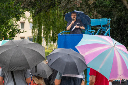 Steffen Wagner steht auf einer erhöhten Bühne unter einem blauen Sonnenschirm und spricht in ein Mikrofon. Im Vordergrund sind mehrere geöffnete Regenschirme zu sehen, darunter ein Regenschirm mit bunten Streifen, vor einer Menschenmenge im Freien.