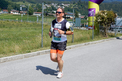 Ein Mann läuft bei einem Marathon auf einer asphaltierten Straße. Er trägt eine Laufnummer am Bauch, eine Sonnenbrille und ein Sportarmband am linken Oberarm. Im Hintergrund sind ein Zaun, grüne Wiesen und Gebäude am Hang zu sehen.