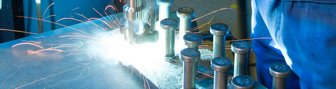 Eine Person in blauer Schutzkleidung führt eine Bolzenschweißung an einer Metallplatte durch. Sichtbar sind Funkenflug, vorbereitete Schweißbolzen und eine Gasflasche im Hintergrund. Die Arbeit erfolgt unter Einsatz einer Bolzenschweißpistole mit Schutzbrille.