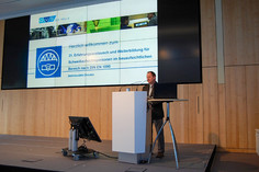 Mann steht an einem Rednerpult in einem modernen Konferenzraum, hinter ihm große Leinwand mit Begrüßungsfolie zur Veranstaltung der SLV Halle und DVS zum Erfahrungsaustausch nach DIN EN 1090