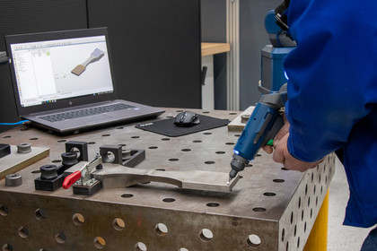 Ein Mann in blauer Arbeitskleidung hält einen portablen 3D-Scanner über ein gebogenes Metallteil auf einem Schweißtisch; auf dem Laptop ist das digitalisierte Werkstück als 3D-Modell sichtbar.