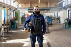 Mann in blauer Arbeitskleidung steht mittig in einer großen, hellen Werkhalle und hält einen Schweißhelm in der Hand, im Hintergrund Werkbänke und technische Arbeitsplätze