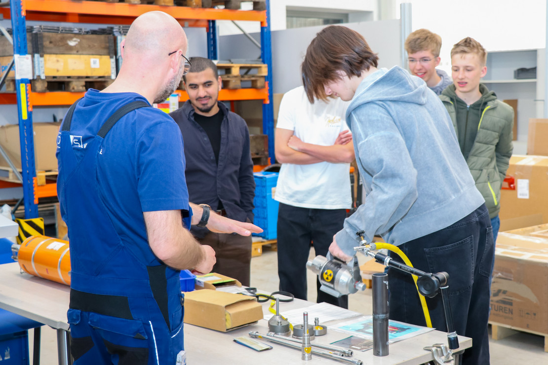 Vier Jungen und ein Mädchen stehen an einem Arbeitstisch mit technischen Bauteilen und Messgeräten in einer Werkhalle. Ein Mann in blauer Arbeitskleidung zeigt auf den Versuchsaufbau, während die anderen Personen den Aufbau betrachten.