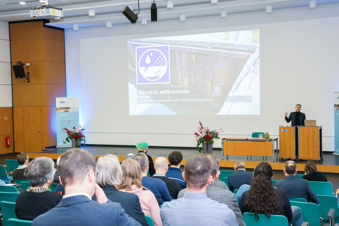 Ein Redner steht an einem Pult in einem Konferenzsaal der SLV Halle und spricht vor einem Publikum. Eine große Präsentationsfolie zeigt den Titel „Herzlich willkommen – 10. Erfahrungsaustausch Korrosionsschutz“.