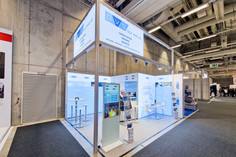 Messestand der SLV Halle mit weiß-blauer Gestaltung, Informationswänden, Stehtischen und einem Prospektständer, umgeben von Betonwänden und Teppichboden in einer Messehalle.