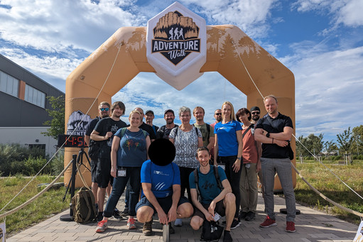 Gruppe posiert vor einem aufblasbaren Tor mit der Aufschrift „ADVENTURE Walk“, eine digitale Uhr zeigt 14:50.