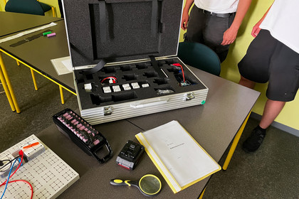Ein offener Aluminiumkoffer mit elektronischen Bauteilen steht auf einem Tisch in einem Schulungsraum, daneben liegen ein Akkupack, eine Bosch-Batterie, eine Lupe und ein Heft mit dem Titel „Batterietechnologie“, im Hintergrund stehen zwei Personen in Arbeitskleidung.