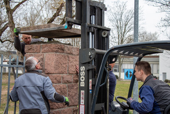 Vier Männer setzen mit einem Gabelstapler eine Steinplatte auf einen gemauerten Pfeiler, einer fährt das Fahrzeug, die anderen positionieren die Platte manuell