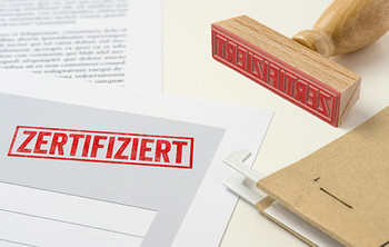 Papierdokument mit rotem Stempelaufdruck „ZERTIFIZIERT“, daneben ein Holzstempel und eine braune Aktenmappe.