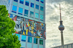 Großflächiges Wandmosaik des ehemaligen „Haus des Lehrers“  zeigt stilisierte Wissenschaftler und Technikmotive; im Hintergrund ragt der Berliner Fernsehturm in den bewölkten Himmel.