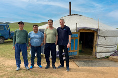 Vier Männer stehen lächelnd vor einer traditionellen mongolischen Jurte auf einer grasbewachsenen Ebene, ein blauer Geländewagen ist im Hintergrund zu sehen.