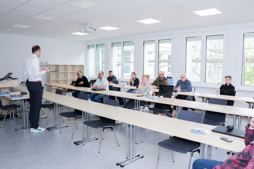 Ein Mann in weißem Hemd hält einen Vortrag vor einer Gruppe von acht Teilnehmenden, die an Tischen in einem Schulungsraum sitzen.