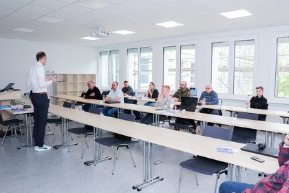 Ein Mann in weißem Hemd hält einen Vortrag vor einer Gruppe von acht Teilnehmenden, die an Tischen in einem Schulungsraum sitzen.