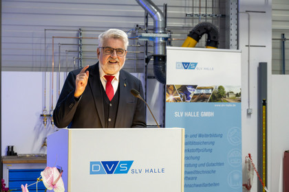 Ein Mann im schwarzen Dreiteiler und roter Krawatte spricht an einem Rednerpult mit DVS SLV Halle-Logo, im Hintergrund befindet sich ein Roll-up mit Leistungsübersicht der SLV Halle GmbH sowie technische Anlagen in einer Werkhalle.