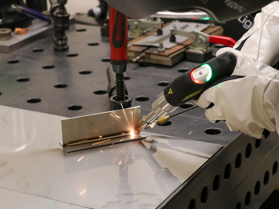 Eine Person mit weißen Schutzhandschuhen bearbeitet ein eingespanntes Metallbauteil mit einem handgeführten Werkzeug, wobei Funken entstehen. Das Werkstück liegt auf einem Arbeitstisch.