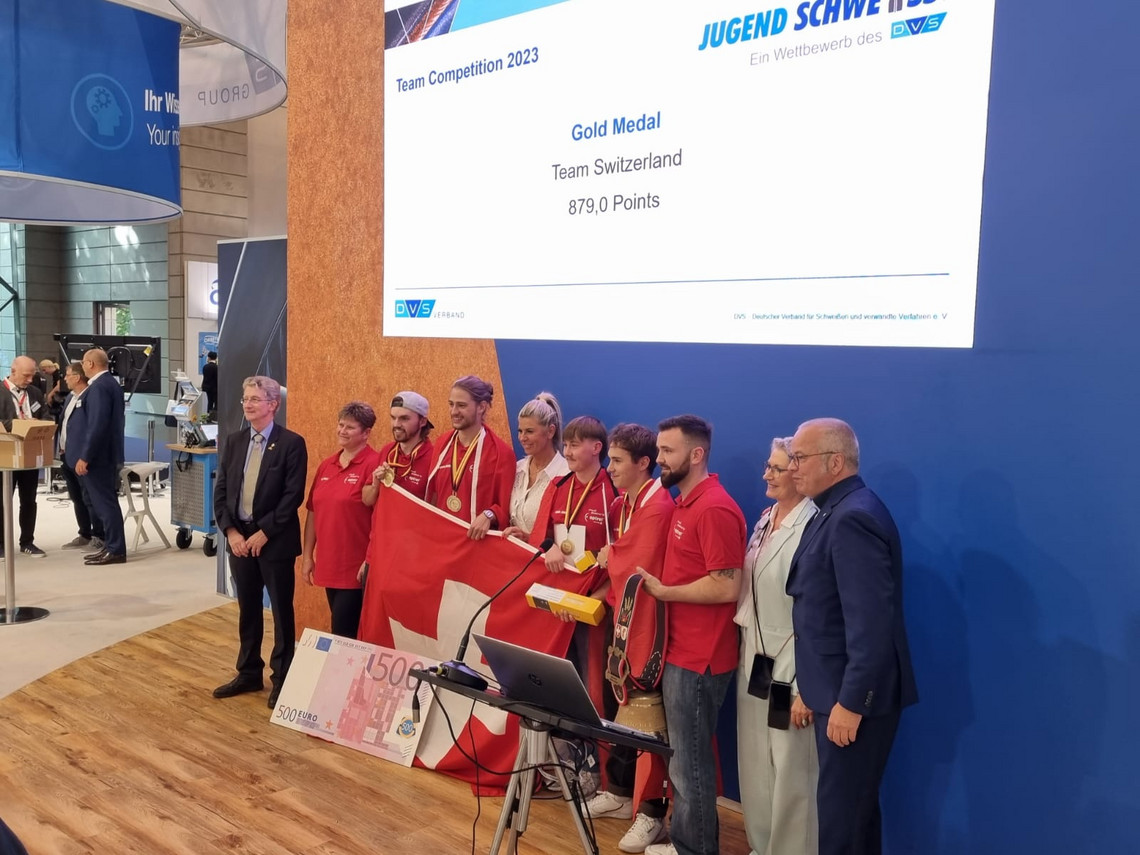 Das Schweizer Gewinnerteam steht mit Medaillen und Landesflagge auf einer Bühne bei der Jugend schweißt Team Competition 2023, ein Display zeigt die Auszeichnung „Gold Medal“ mit 879,0 Punkten.