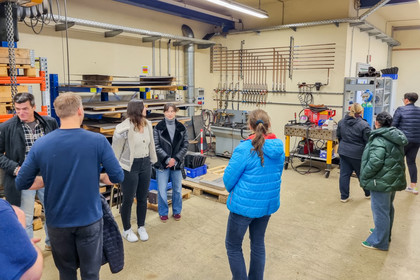 Mehrere Personen stehen in einer Werkstatt und unterhalten sich zwischen Werkbänken und Maschinen über technische Arbeitsprozesse.