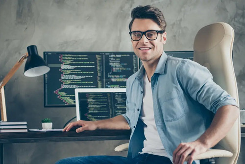 Junger Mann mit Brille sitzt lächelnd am Schreibtisch vor mehreren Monitoren mit Programmcode, sinnbildlich für Softwareentwicklung und IT-Arbeit.