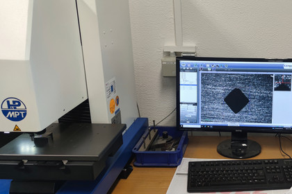 Ein automatisiertes Härteprüfgerät von MPT steht auf einem Arbeitstisch neben einem Monitor, der eine stark vergrößerte Prüfeinprägung in Form eines Rhombus auf einer Metalloberfläche anzeigt. Daneben liegen Werkzeuge und eine Tastatur.