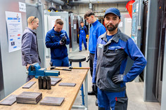 Wettbewerbssituation in der Werkhalle mit mehreren Teilnehmenden in blauer Arbeitskleidung, im Vordergrund Bauteile und Werkzeuge, eine Fachkraft blickt offen und freundlich in die Kamera