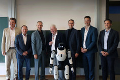 Sechs Männer in Anzügen stehen für ein Gruppenfoto  in einem Raum mit einer großen Projektionsfläche, davor ein weiß-schwarzer humanoiden Roboter.