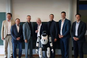 Sechs Männer in Anzügen stehen für ein Gruppenfoto  in einem Raum mit einer großen Projektionsfläche, davor ein weiß-schwarzer humanoiden Roboter.
