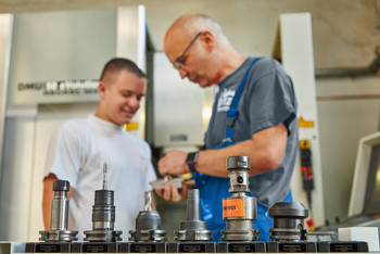 Ein Ausbilder erklärt einem jungen Teilnehmenden in einer Werkstatt die Handhabung verschiedener Fräswerkzeuge, im Vordergrund liegen mehrere Werkzeugaufnahmen © SLV Halle GmbH 2025