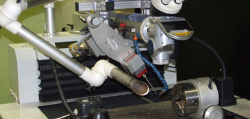 Festkörperlaser: Ein Laserschweißsystem mit integriertem Mikroskop und Roboterarm ist auf einem metallischen Arbeitstisch montiert. Das zu bearbeitende Bauteil ist in einer drehbaren Spannvorrichtung eingespannt, daneben befinden sich Präzisionswerkzeuge und ein Steuerdisplay.