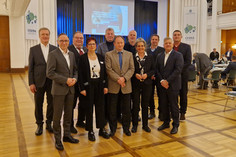 Elf Personen, acht Männer und drei Frauen, stehen in einem festlich beleuchteten Saal in zwei Reihen und blicken in die Kamera, im Hintergrund Bühne mit Präsentation und Tische mit Teilnehmenden