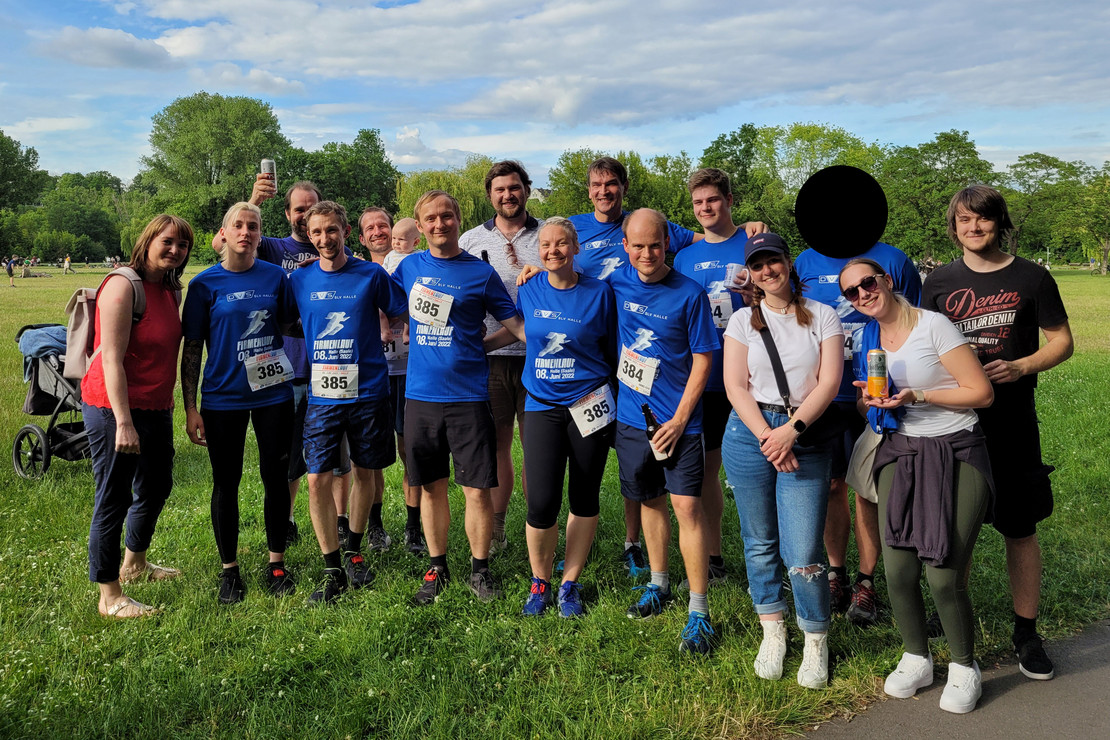 Gruppe von 16 Personen (10 Männer, 6 Frauen) posiert nach einem Firmenlauf im Park, einige tragen blaue Laufshirts mit Startnummern, andere Freizeitkleidung, im Hintergrund grüne Wiese und Bäume.