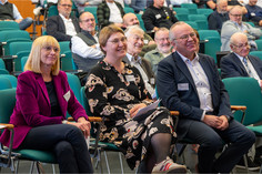 Drei Personen sitzen in der ersten Reihe eines Vortragssaals. Eine Frau in violettem Blazer, eine Frau im gemusterten Kleid und ein Mann im dunklen Sakko hören aufmerksam zu, hinter ihnen sitzen weitere Teilnehmende.