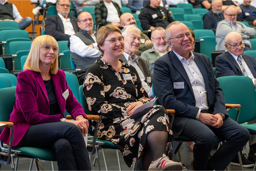 Drei Personen sitzen in der ersten Reihe eines Vortragssaals. Eine Frau in violettem Blazer, eine Frau im gemusterten Kleid und ein Mann im dunklen Sakko hören aufmerksam zu, hinter ihnen sitzen weitere Teilnehmende.