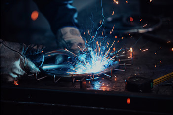 Löter mit Schutzhandschuhen bearbeitet ein rundes Metallteil, dabei sprühen helle Funken und blaues Licht auf der Werkbank.