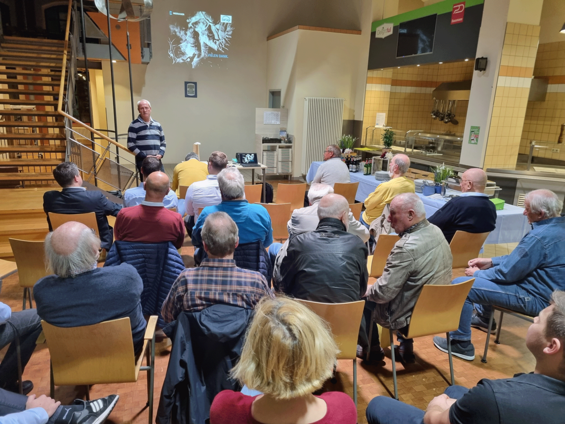 Ein Mann steht vor einer sitzenden Zuhörergruppe und hält einen Vortrag, im Hintergrund läuft eine Präsentation mit einem Schweißmotiv auf der Wand; die Veranstaltung findet in einem Saal mit Treppe, Fliesenbereich und Holzboden statt.