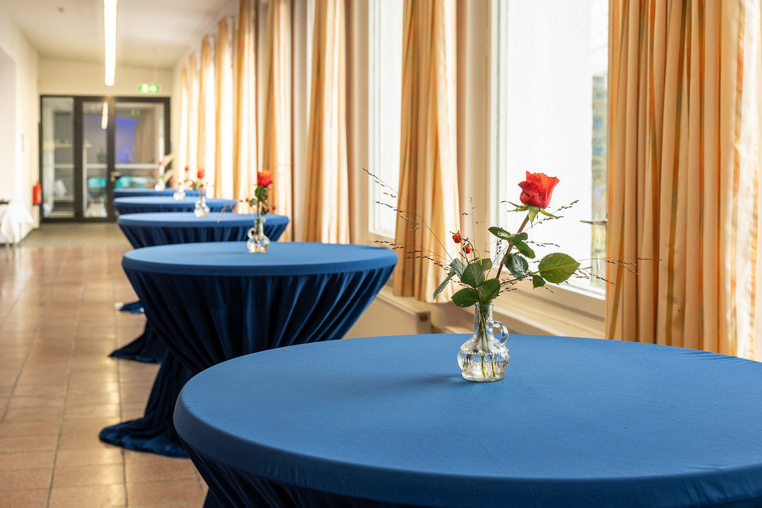 Reihe von runden Stehtischen mit blauer Tischdecke und einzelnen roten Rosen in Vasen, entlang eines lichtdurchfluteten Flurs mit großen Fenstern.