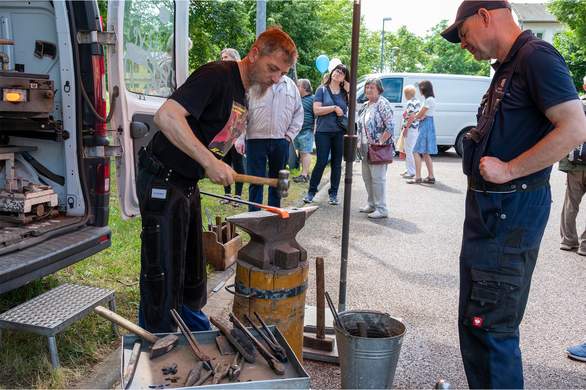 Drei Männer stehen an einem mobilen Arbeitsstand im Freien und beobachten, wie ein Schmied ein eingespanntes Metallstück bearbeitet.