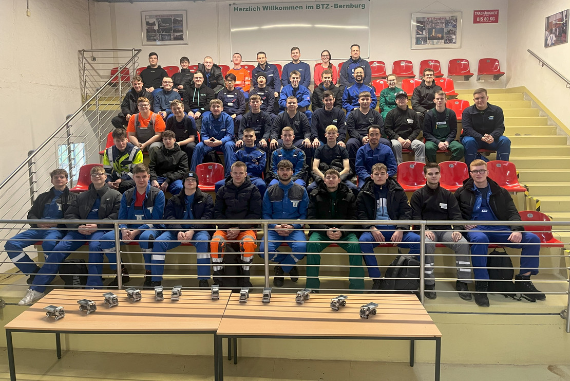 Gruppenfoto mit rund 50 Auszubildenden auf Tribünenstufen im BTZ Bernburg, vorrangig männlich besetzt; alle tragen Arbeitskleidung in verschiedenen Farben, vorne auf Tischen liegen Werkstücke.